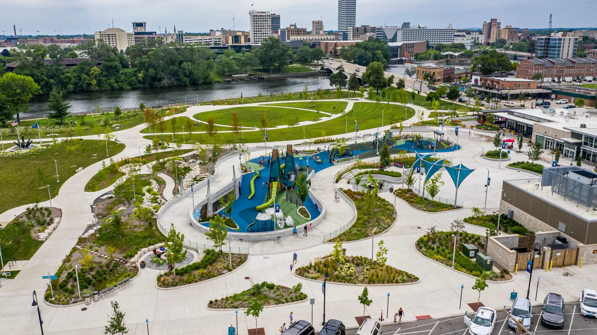 An aerial view of a modern public park featuring a blue, winding playground area, green landscaping, and a river backdrop.