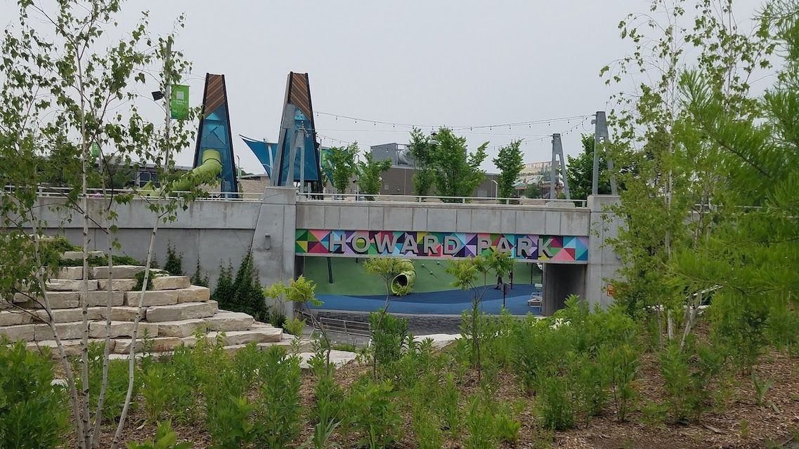 A concrete pedestrian bridge with a colorful, geometric mural, set in a park with terraced stone stairs and trees.