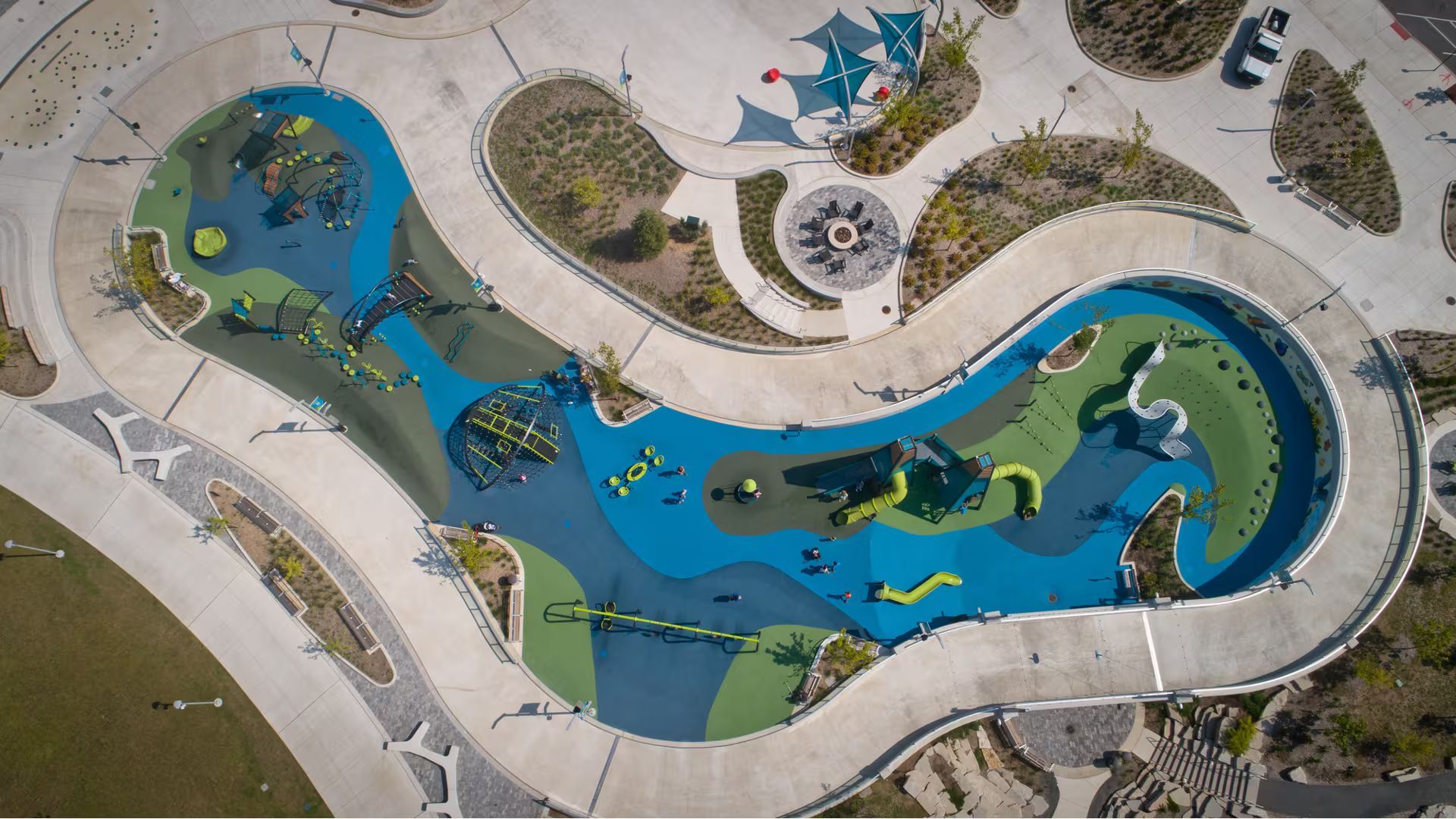 An aerial view of a colorful, winding outdoor playground with blue and green ground surfaces surrounded by a concrete path.
