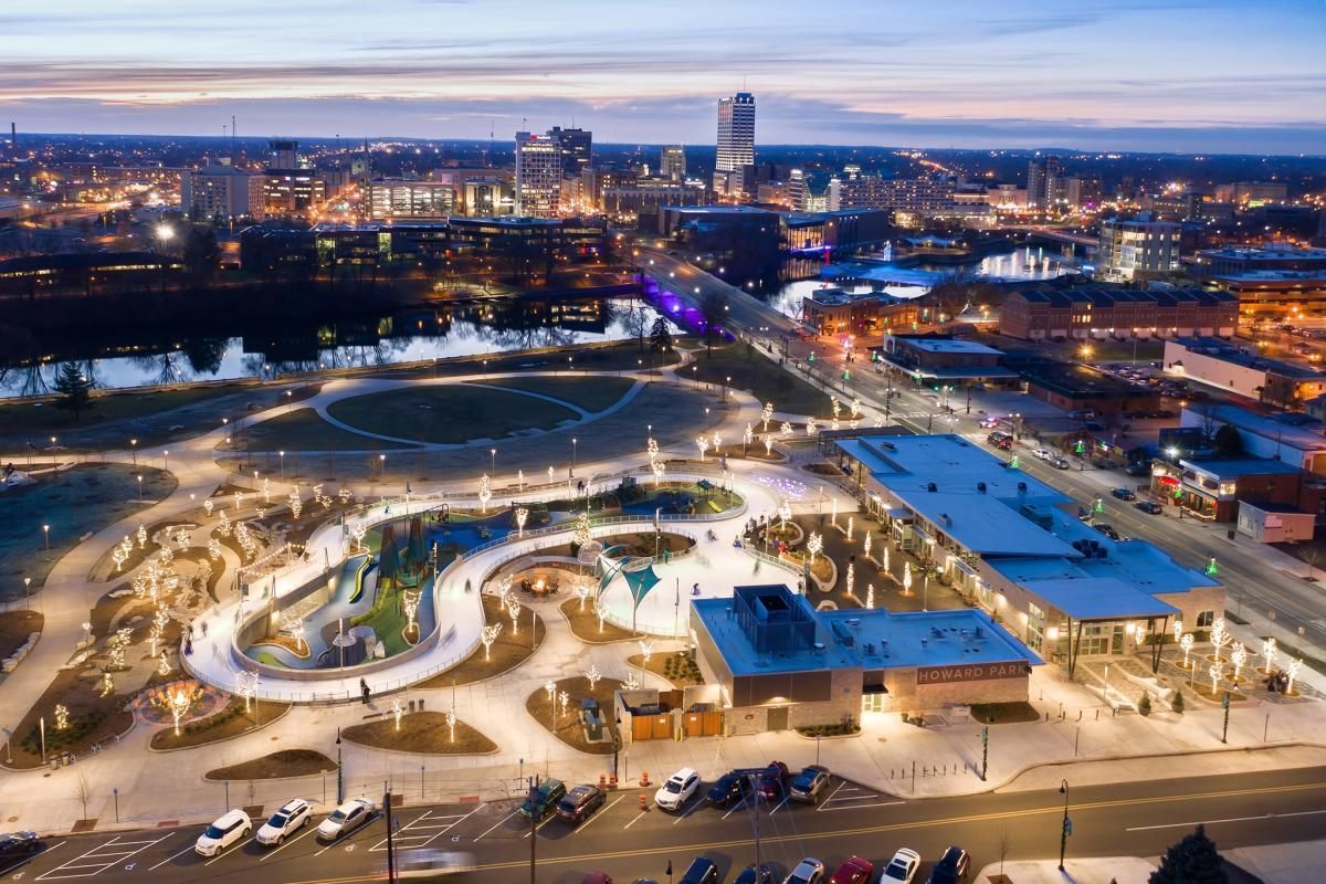 An aerial night view of a glowing ice skating trail and park facility in a city with a skyline in the background.
