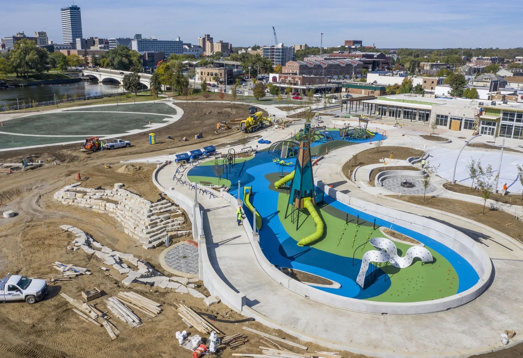 An aerial view of a colorful, modern playground under construction near a river and city skyline.