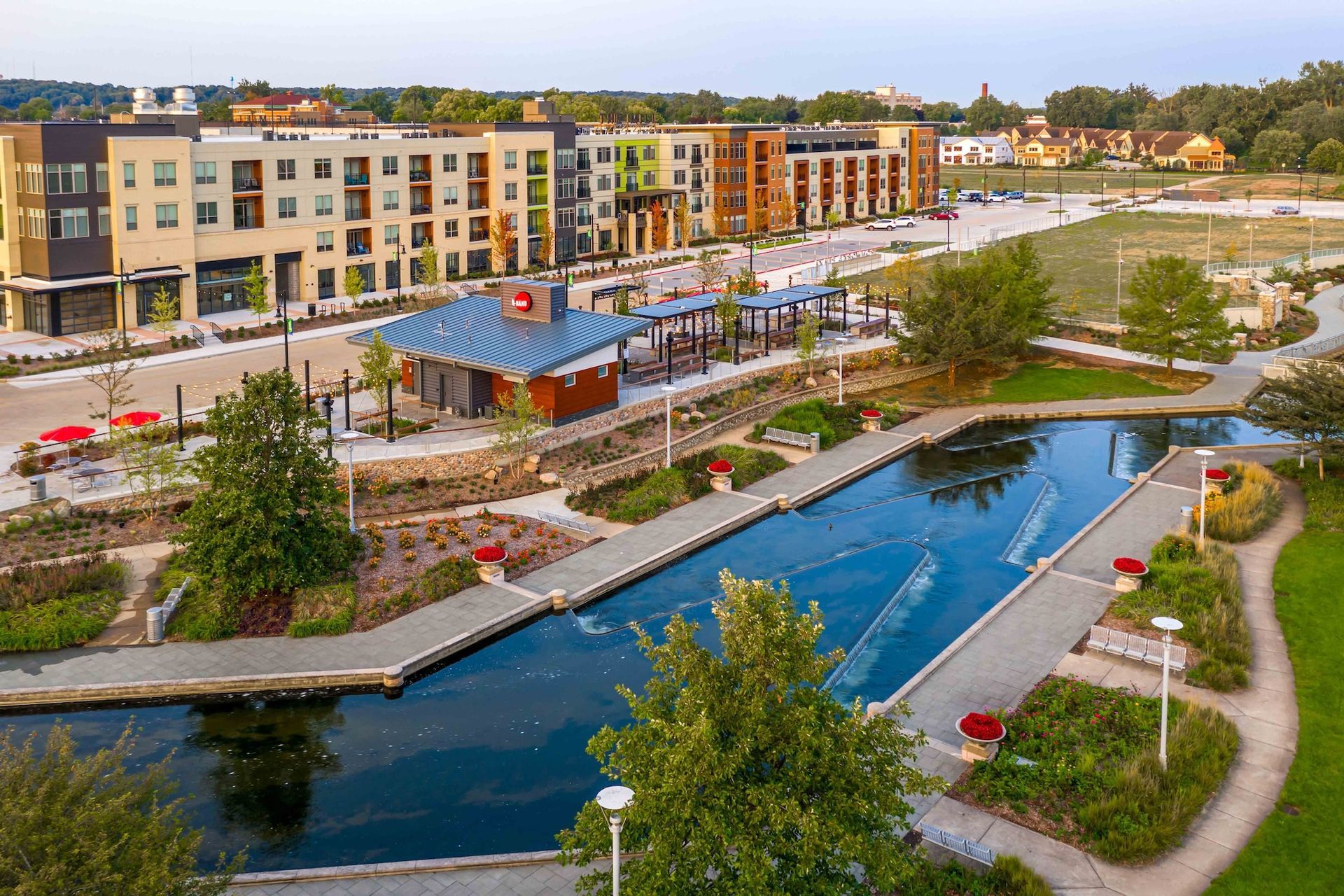 An aerial view of a modern multi-story apartment building bordering a rectangular pond with walkways and landscaping.