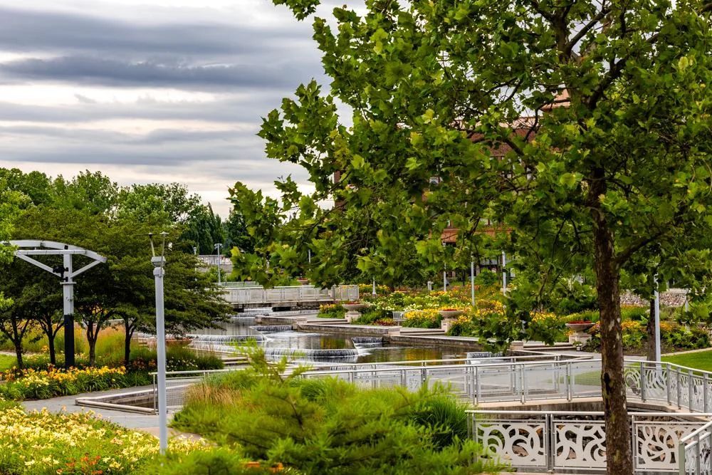 Lush trees and vibrant flower beds frame a peaceful, multi-level water fountain in a park under a cloudy sky.