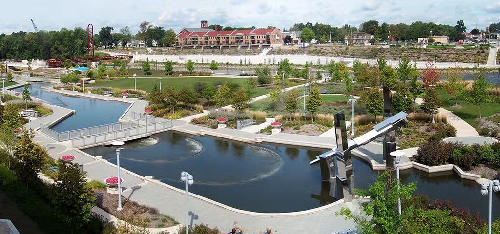 An elevated view of a landscaped city park with a central pond, walking paths, gardens, and distant buildings.