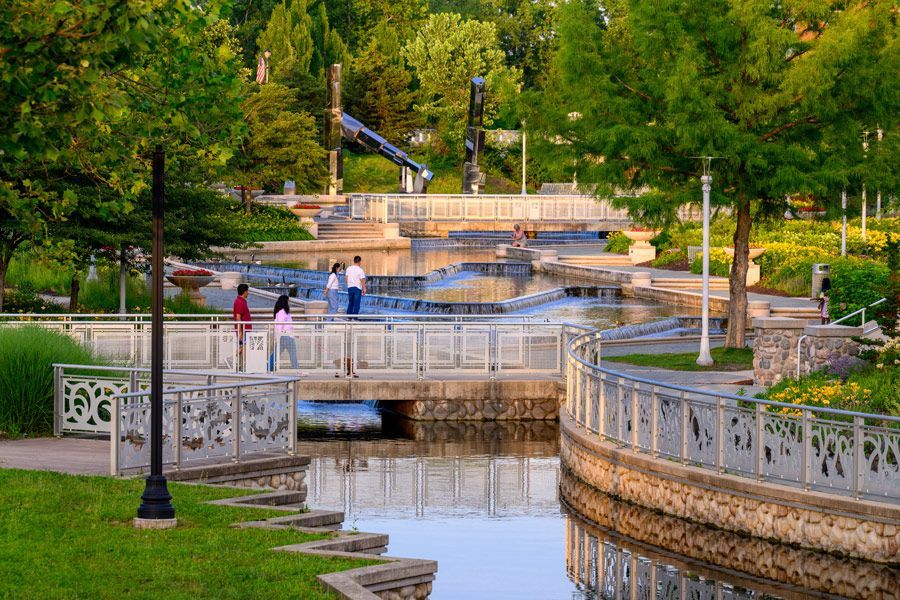 A sunlit public park with a stone-lined canal, decorative metal railings, a pedestrian bridge, and lush green trees.