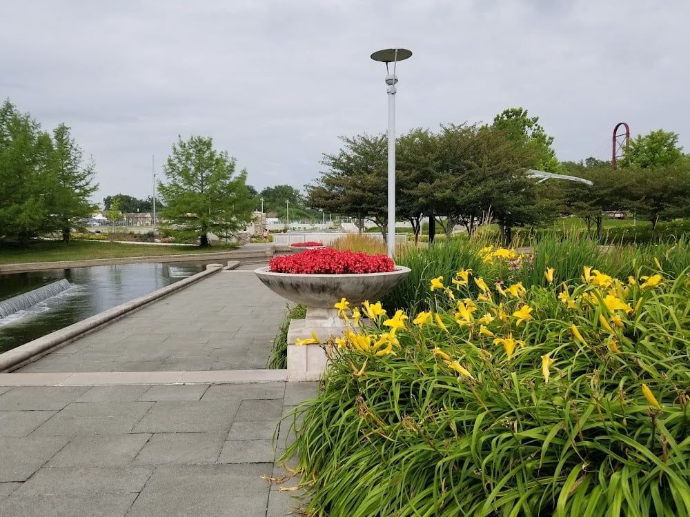 A stone path leads past a circular planter of red flowers and a bed of yellow daylilies toward a park pond.