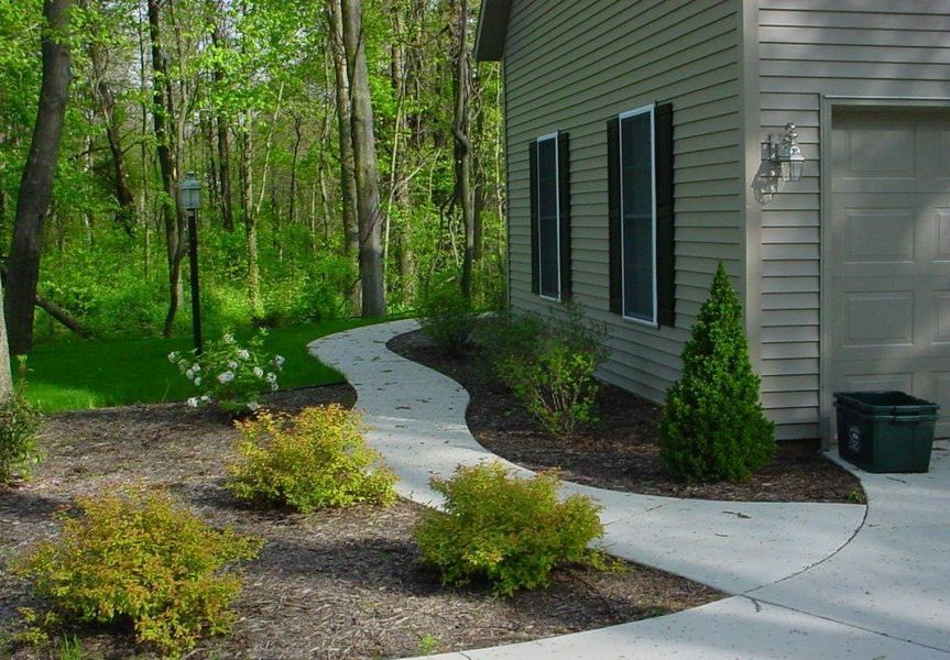 Curving concrete path leads from a driveway towards a house, lined with bushes and trees.