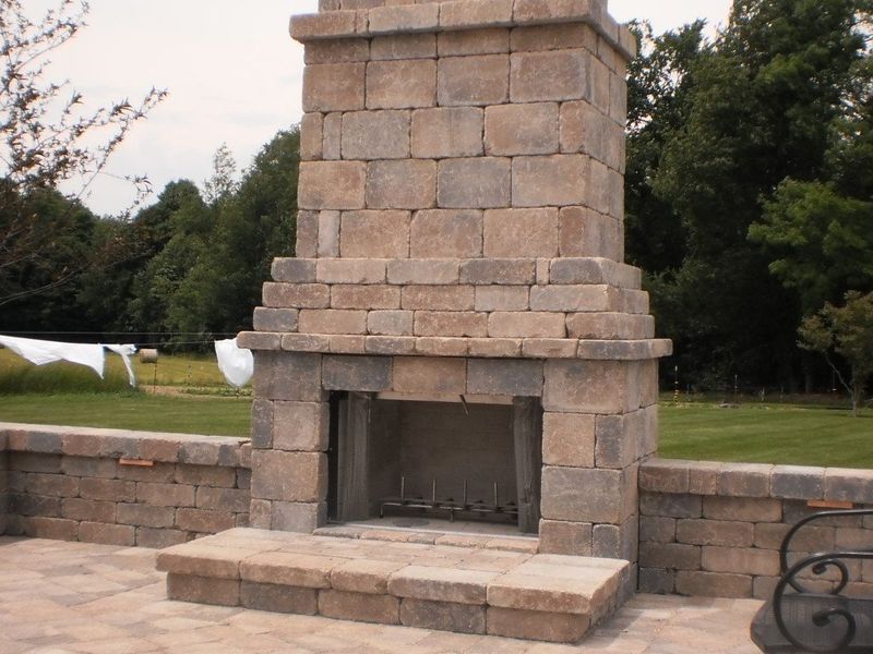 Stone outdoor fireplace with a brick patio, set against a lawn and trees.