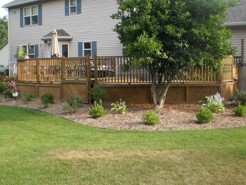 Backyard deck with tree, surrounded by mulch and shrubs; house in background with grass lawn.