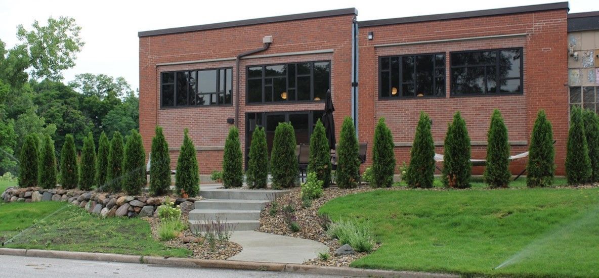 Brick building with large windows and a row of evergreen trees along the front. A short stone wall 