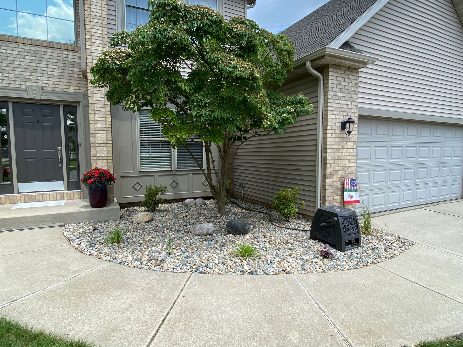 A landscaped island with a tree and rocks sits between a tan brick home's entryway and its garage on a concrete driveway.