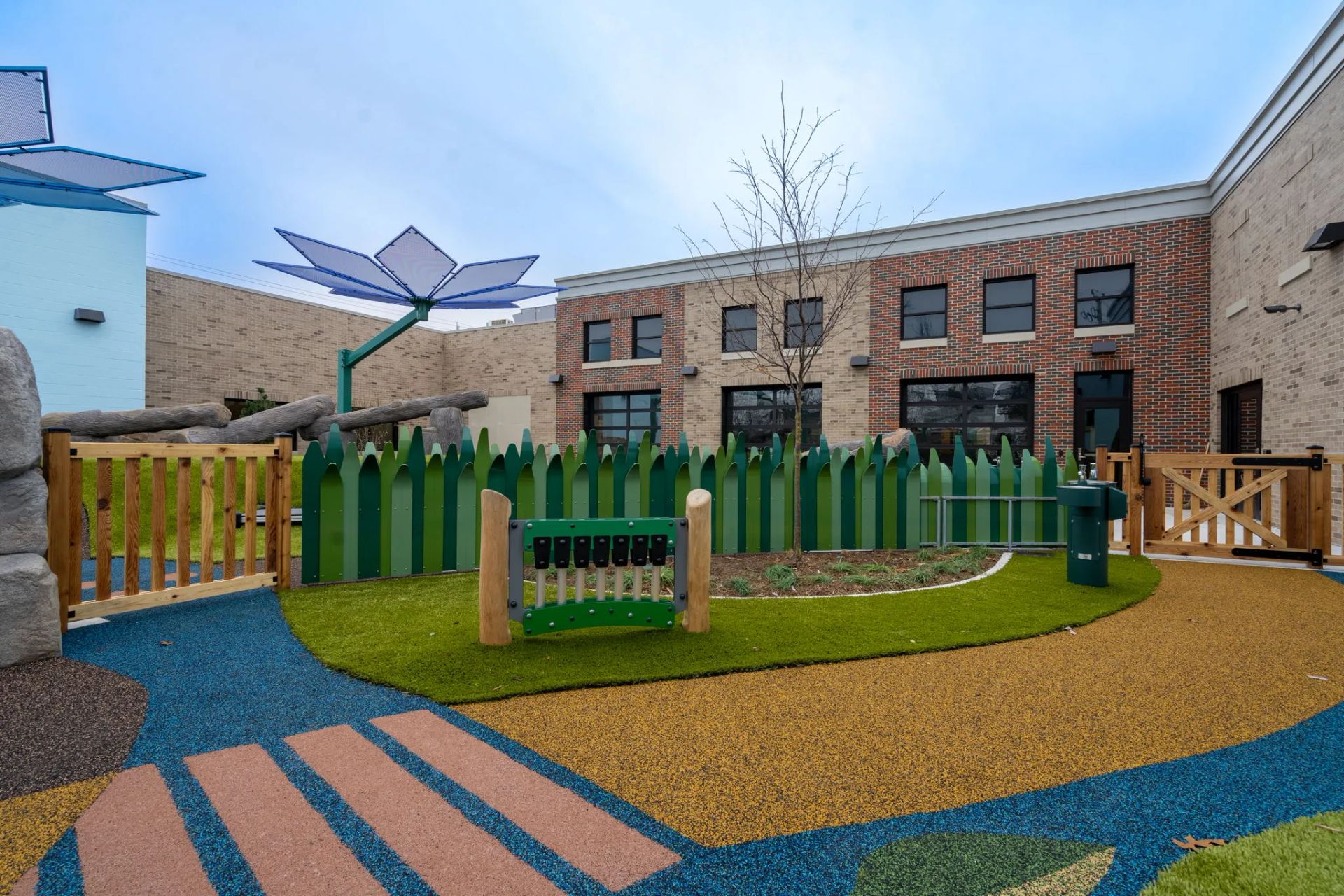A colorful, fenced-in outdoor playground with rubberized surfacing, artificial grass, and a flower-shaped shade structure.