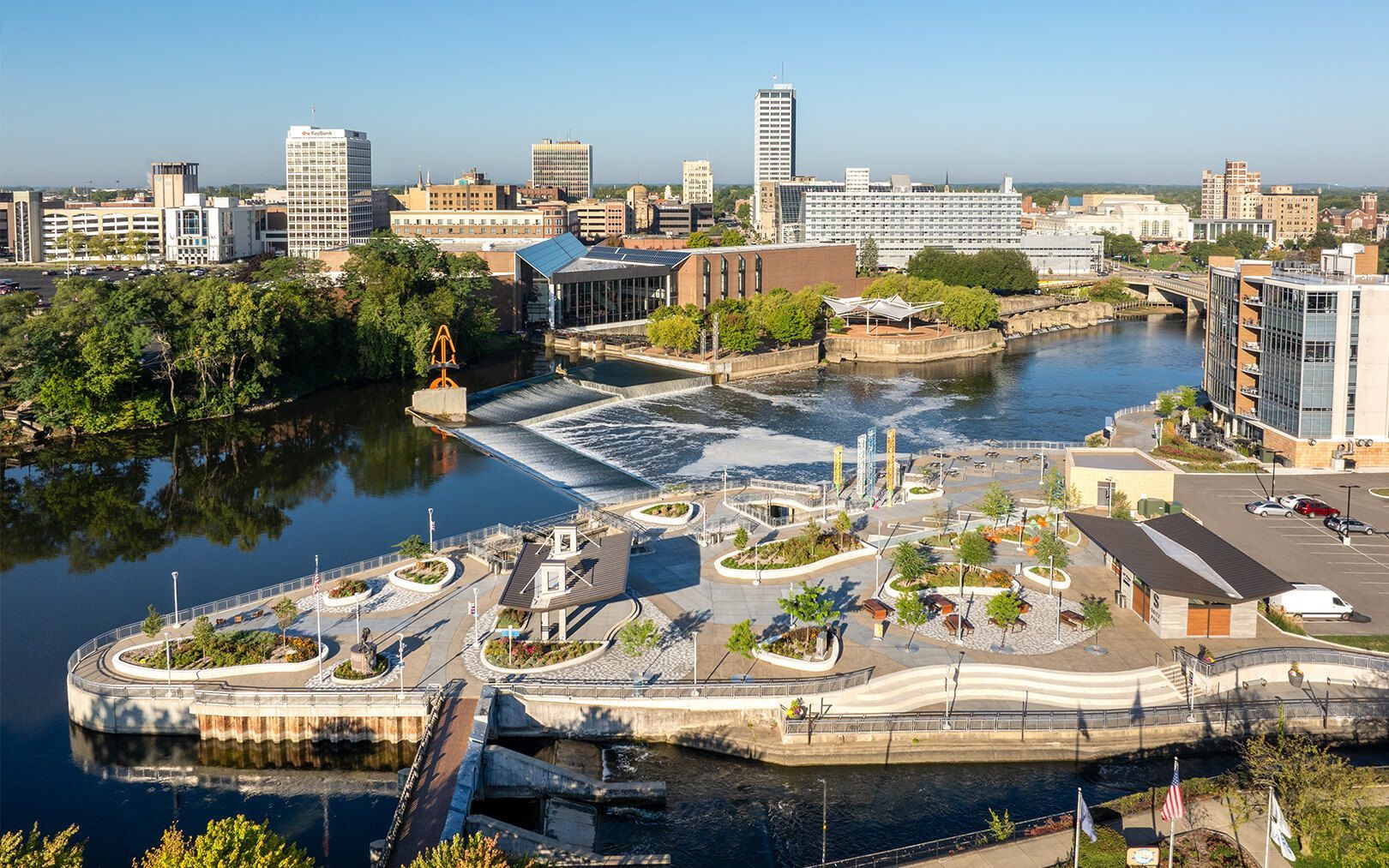 An aerial view of a riverside park with walking paths, landscaping, and a small waterfall, set against a city skyline.