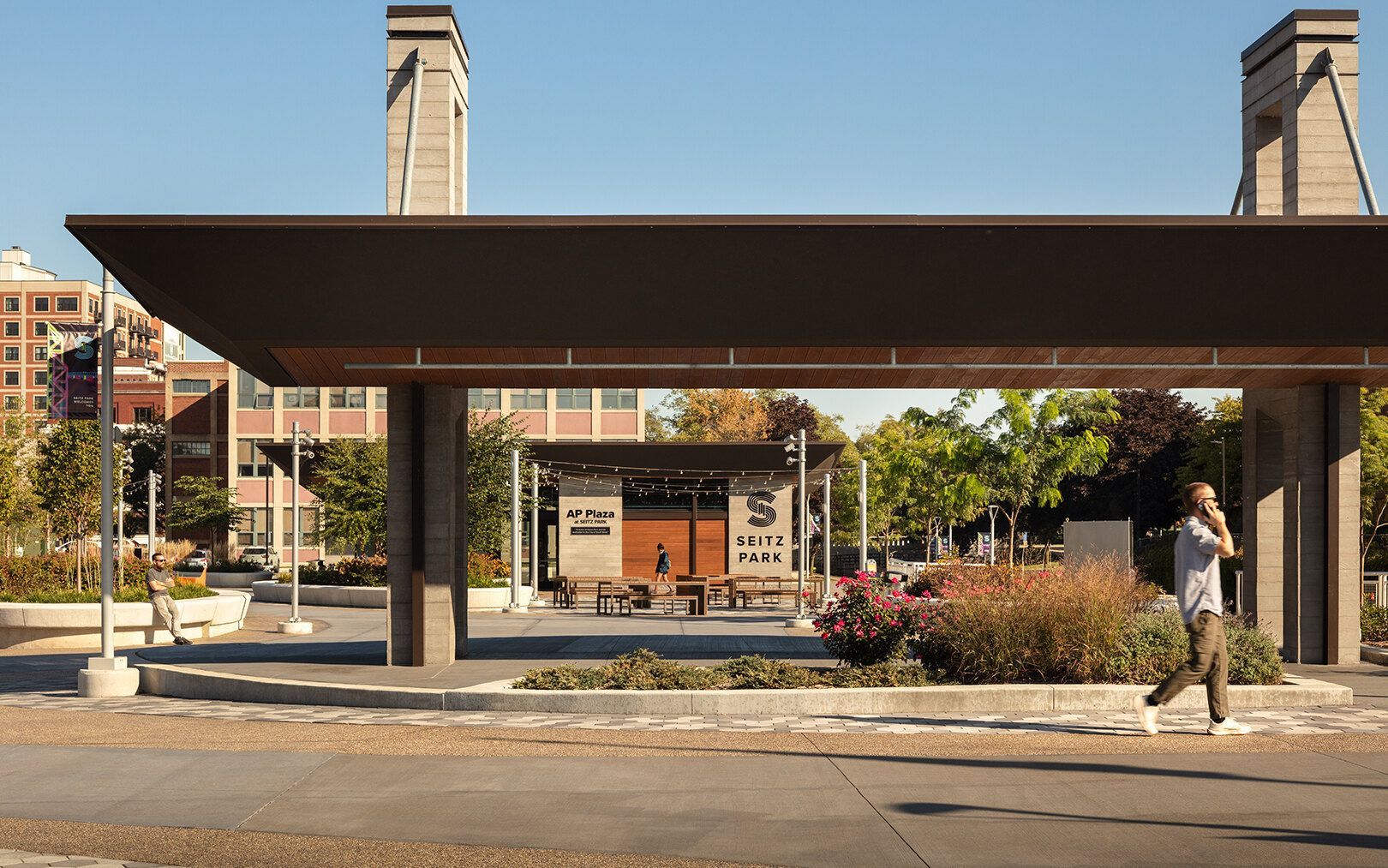 A person walks past a modern outdoor pavilion with stone pillars, wooden accents, and surrounding landscaping.