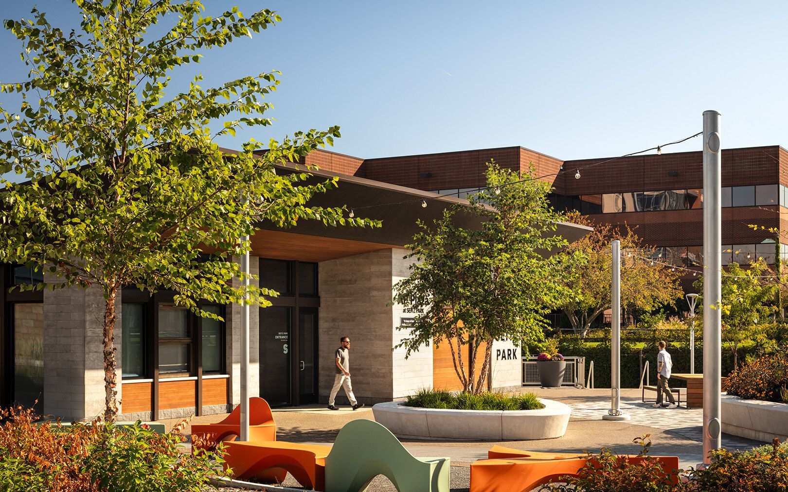 An outdoor courtyard with orange bench seating, trees, and people walking near a modern brick and stone building.
