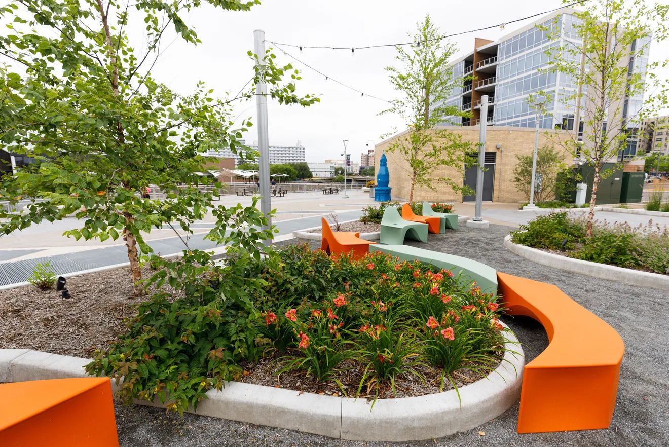 Modern urban park with orange curved benches, small trees, and a flower bed on a gravel path near a building.