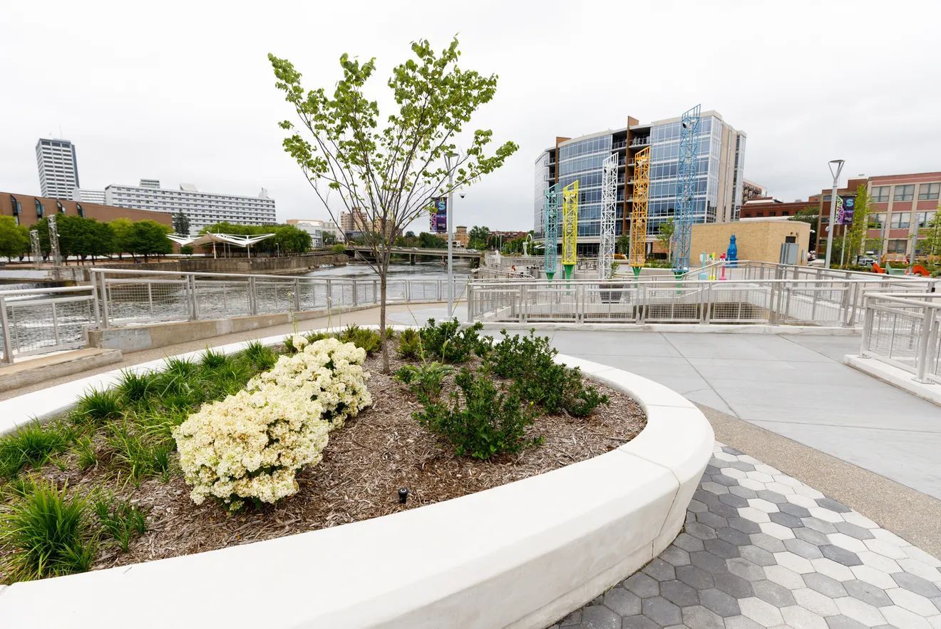 A curved white concrete planter with shrubs and a small tree overlooks a riverfront plaza with modern glass buildings.