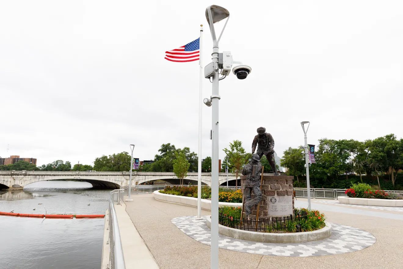 A bronze monument of two figures stands on a riverside path near an American flag, a bridge, and a security camera pole.