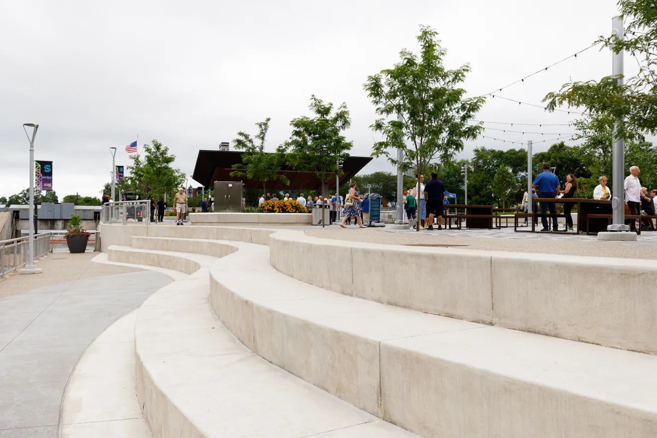Curved concrete steps serve as amphitheater seating in a park with trees, string lights, and people in the background.
