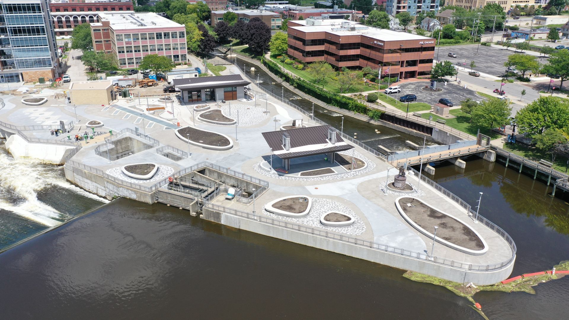 An aerial view of a riverfront park with concrete walkways, landscaping, and adjacent office buildings.