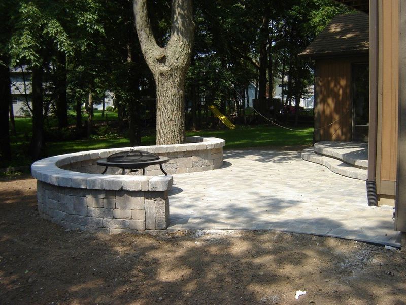 Brick patio with curved retaining wall and fire pit. Mature tree in the background, next to a gazebo.