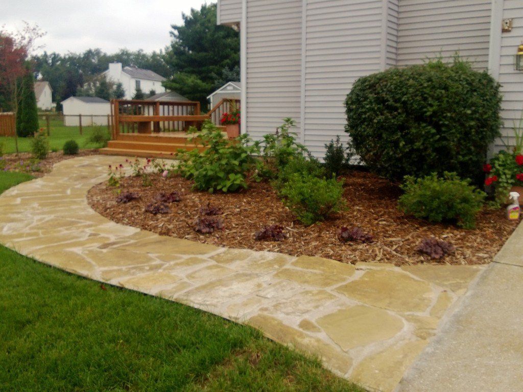Stone pathway curves toward a house with a wooden deck; landscaped bed with mulch, shrubs, and flowers.
