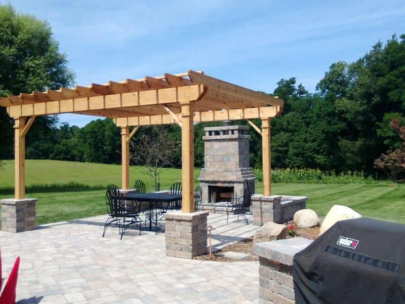 Patio with wooden pergola, fireplace, and seating on a brick surface, surrounded by greenery.