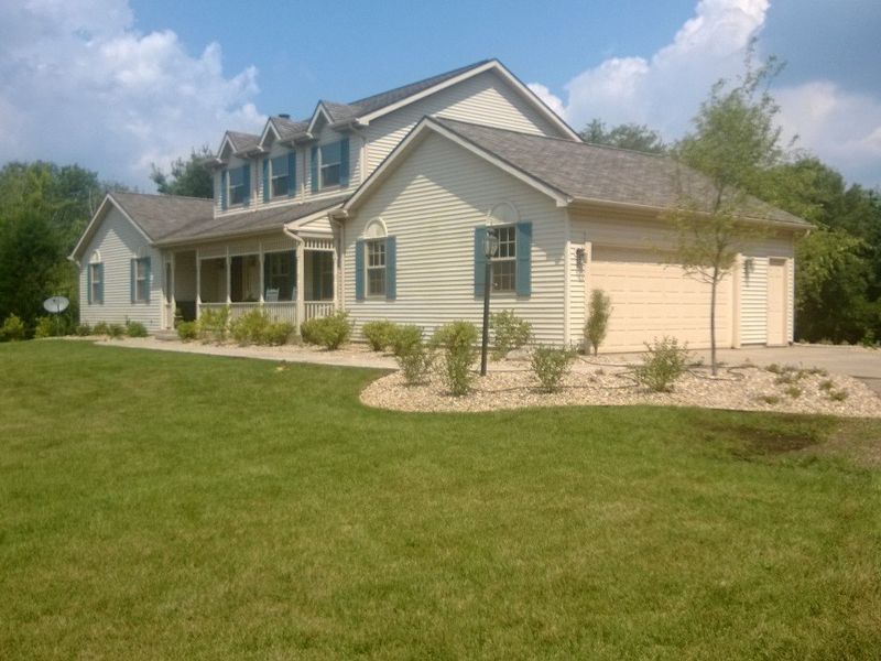 Light-colored two-story house with green shutters, a porch, and a two-car garage, on a grassy lawn with a blue sky.