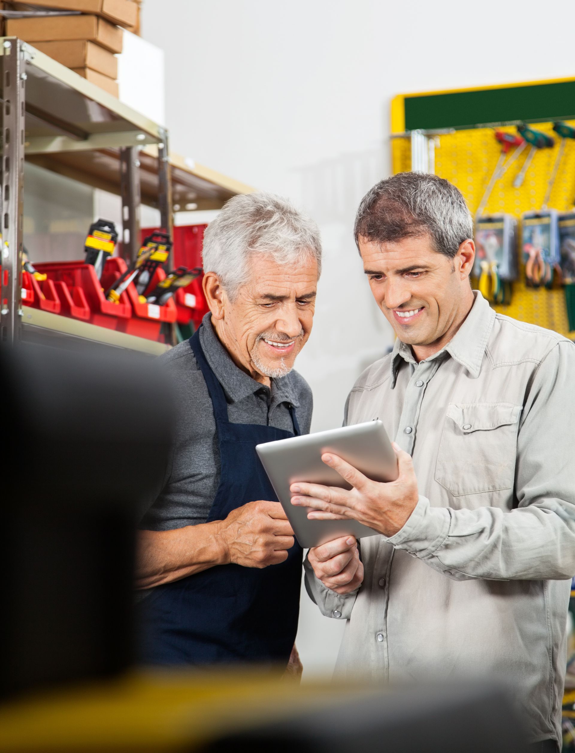 Two workers in a warehouse reviewing a tablet together, with shelves and tools in the background