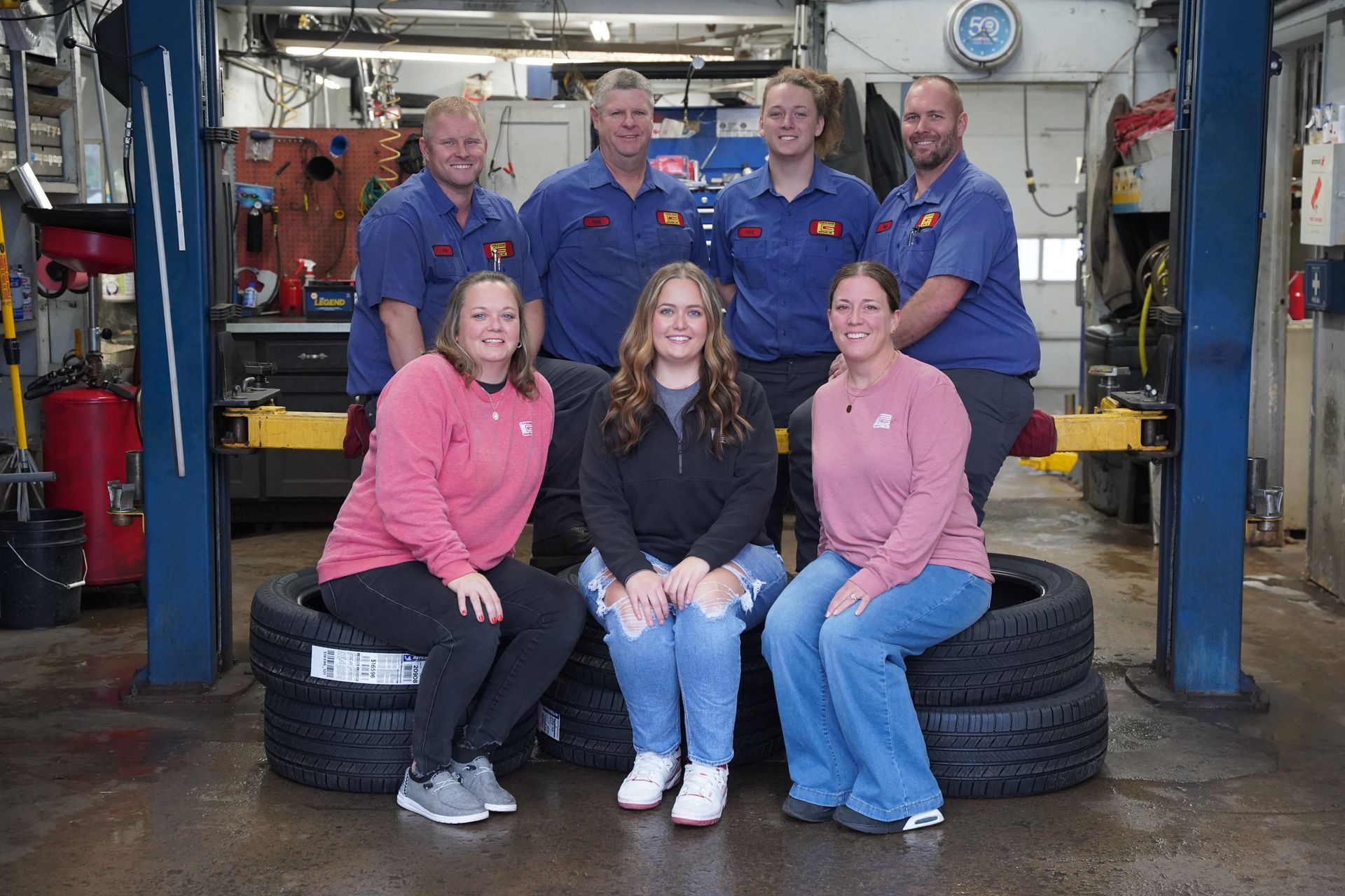 Six mechanics in blue and pink work uniforms posing in an auto repair garage, with tires and a car lift behind them