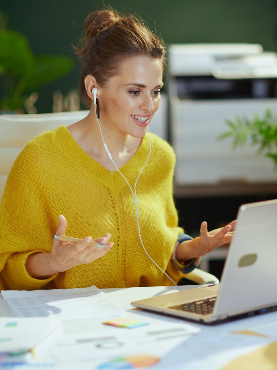 Woman in yellow sweater video chatting at a desk with a laptop and papers spread out.