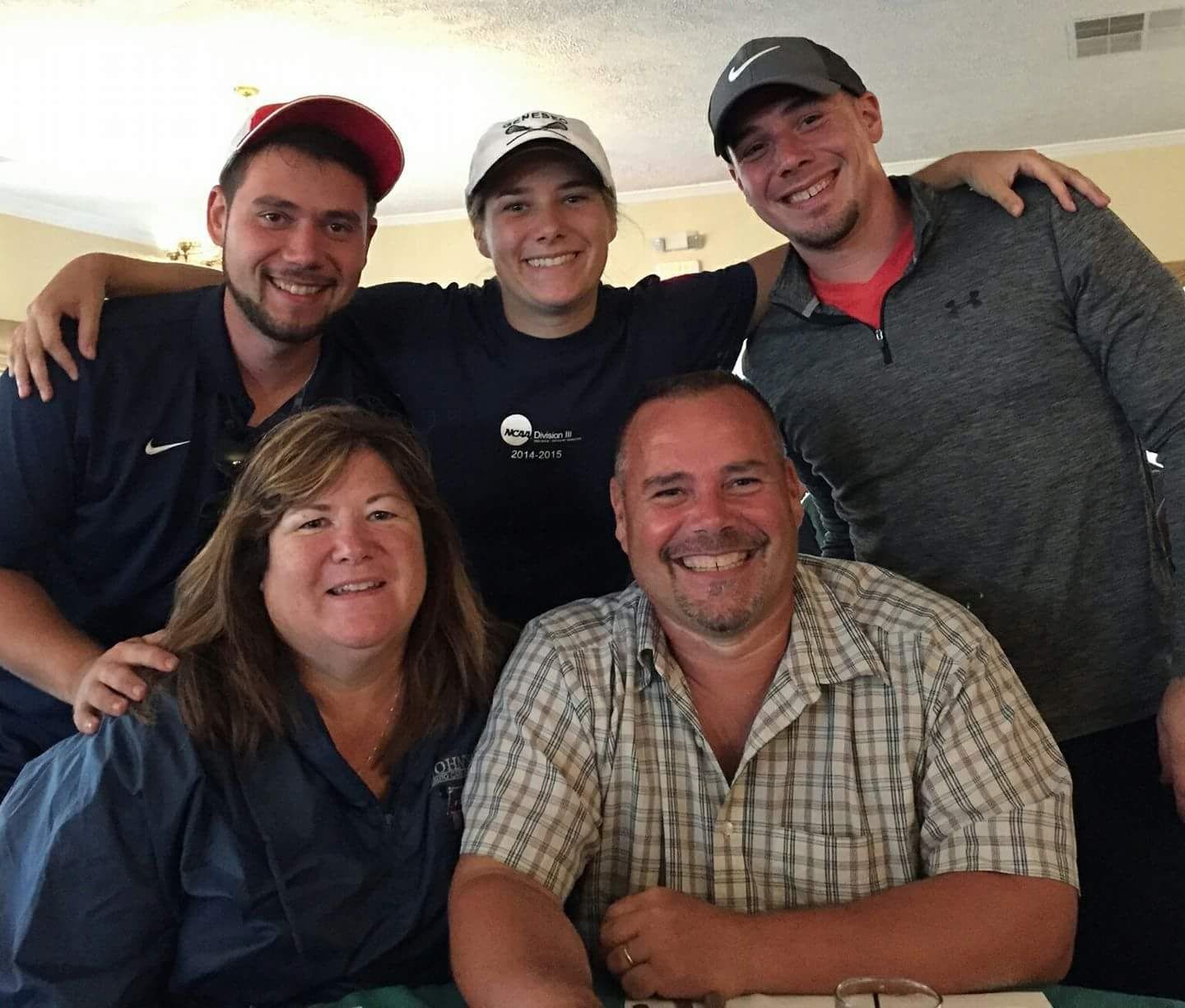 Five smiling people posing together indoors, with two seated in front and three standing behind.