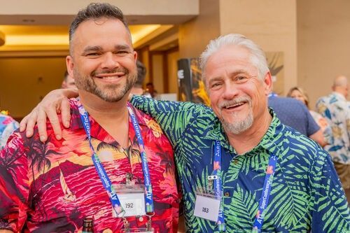 Two smiling men with conference badges, standing arm-in-arm in colorful tropical shirts.