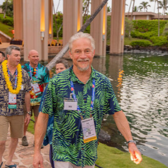 Smiling man in a blue tropical shirt walking by a pond with others and palm trees nearby