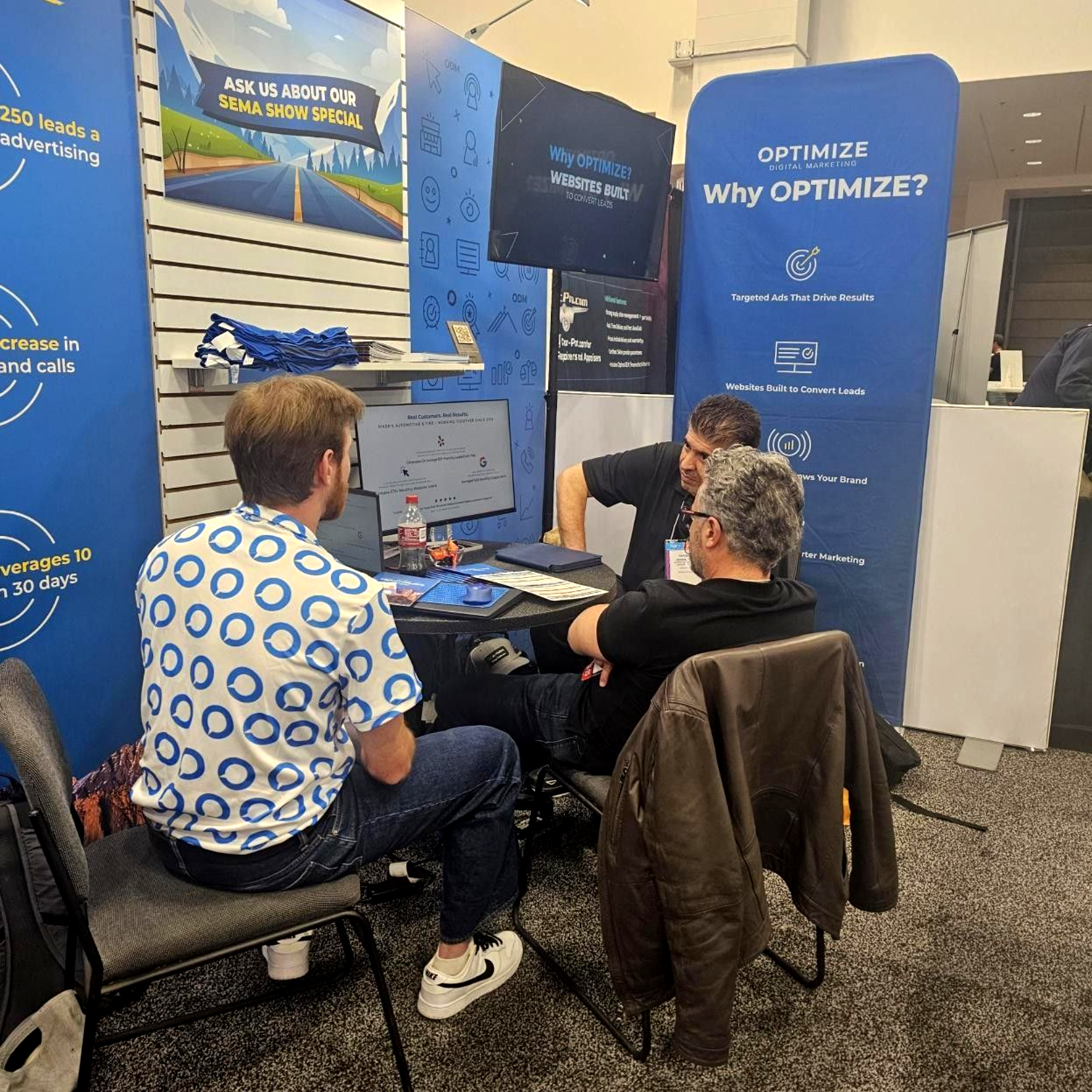 Three people talking at a trade show booth with blue Optimize posters and a laptop on the table