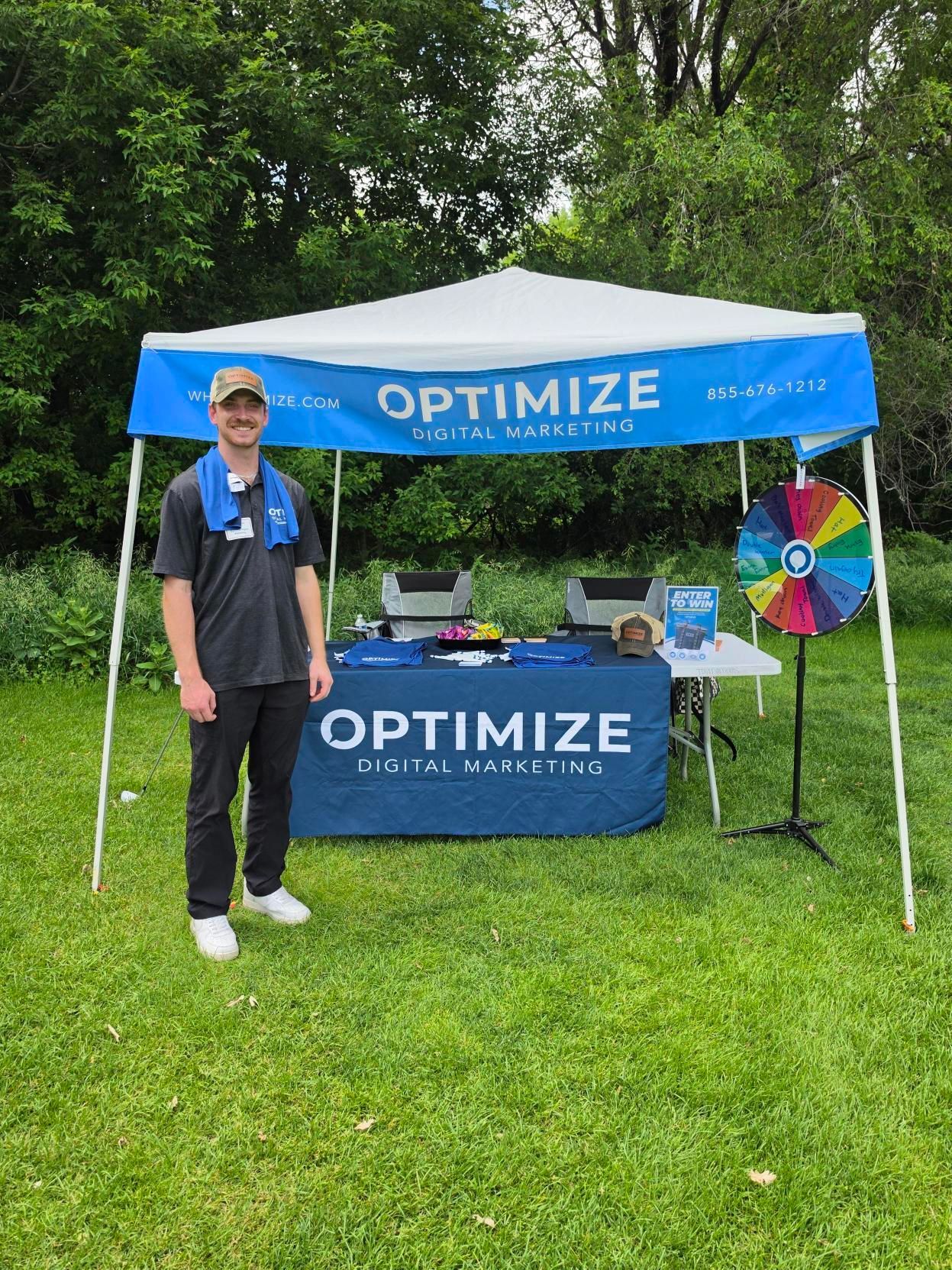 Person at an OPTIMIZE booth with blue canopy, table, and colorful wheel in a grassy park