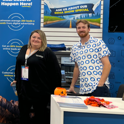 Two booth staff at a trade show table with a “Sema Show Special” banner and promotional displays
