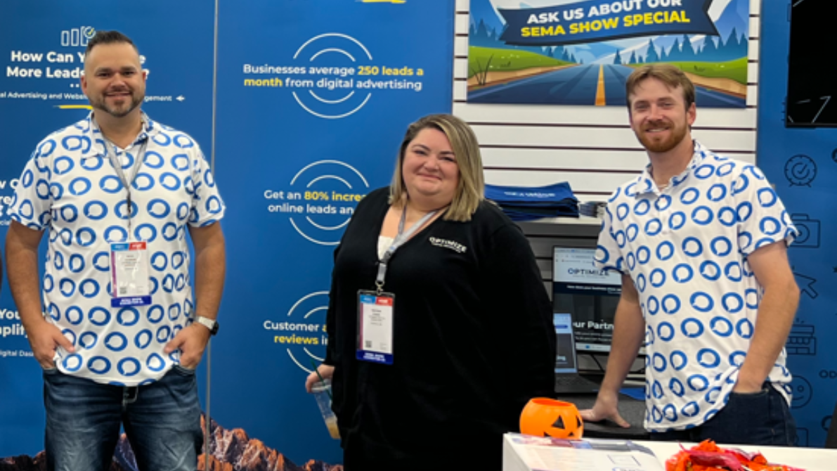 Three people posing at a trade booth with blue banners and branded shirts.