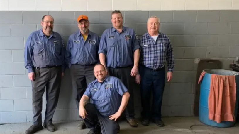 Five people pose in blue shirts against a gray wall, one kneeling in front, with a striped table at right.