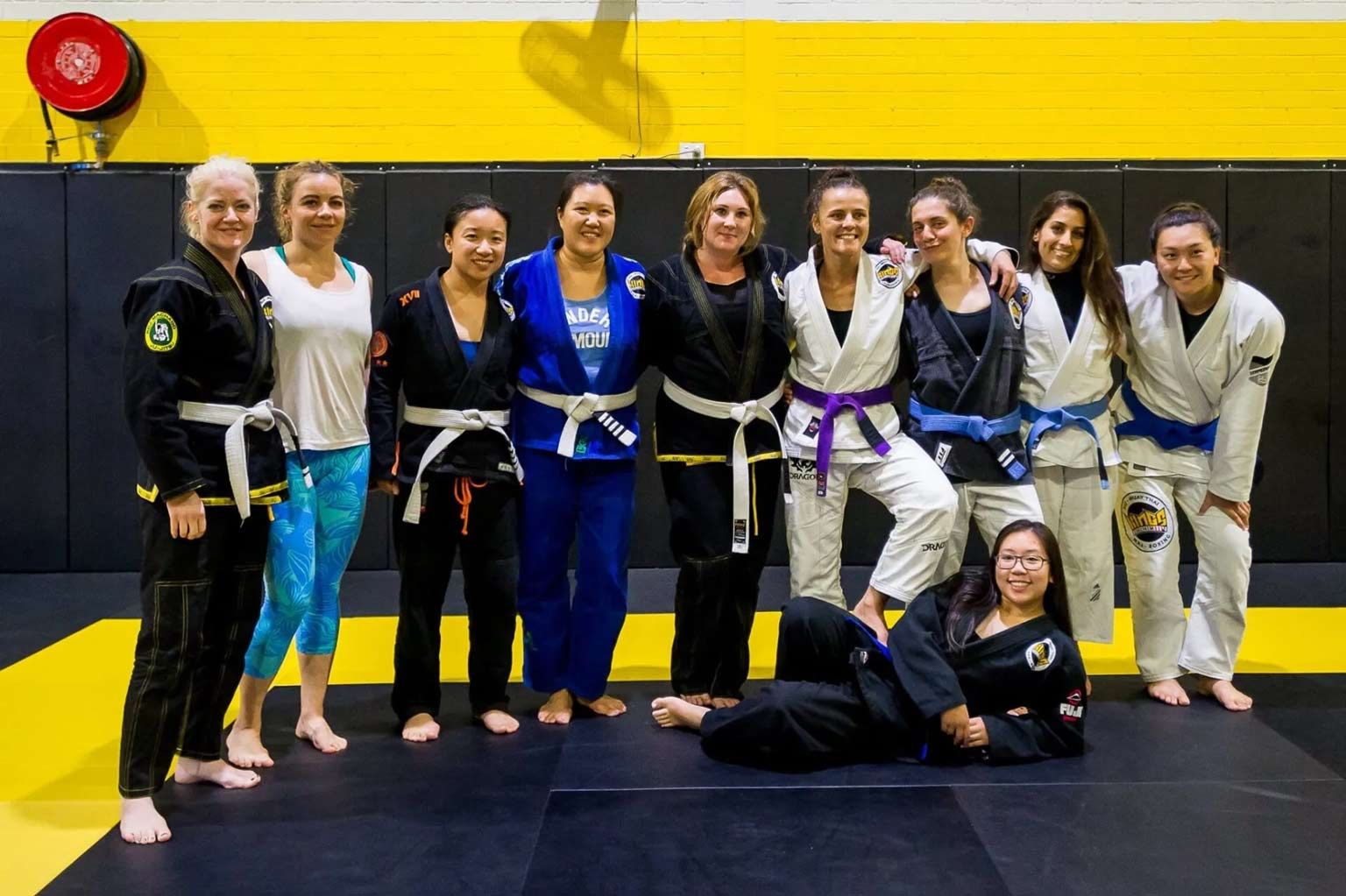 Group of people in jiu-jitsu gis posing on a mat in a gym, with one person kneeling in front.