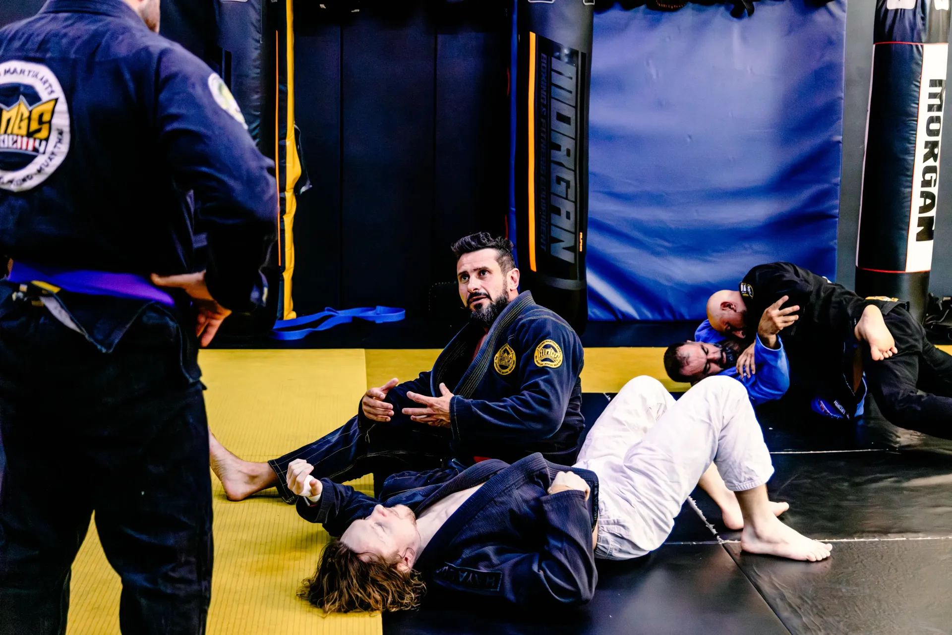 People training Brazilian jiu-jitsu on mats in a gym, wearing gis and rash guards