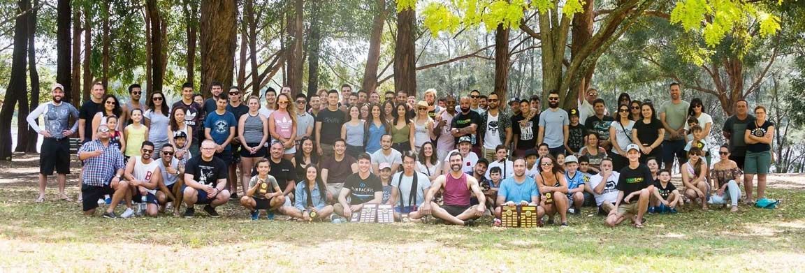 Large group posing together outdoors among trees in a sunny wooded area