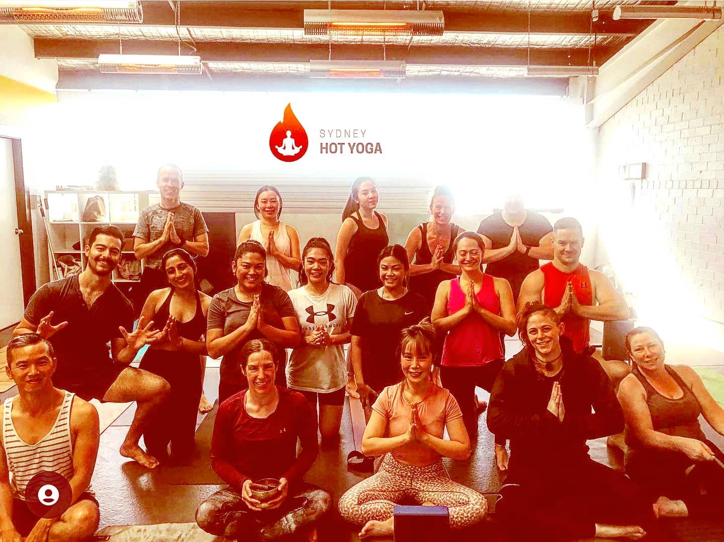 Group yoga class in a bright studio, seated in meditation pose with hands together, under a “HOT YOGA” sign.