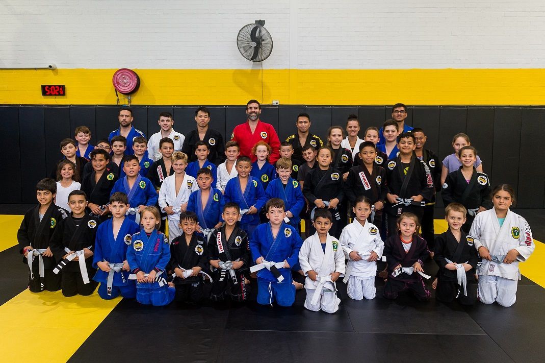 A large group in gi pose for a photo in a BJJ gym with mats, posters, and gym equipment behind.