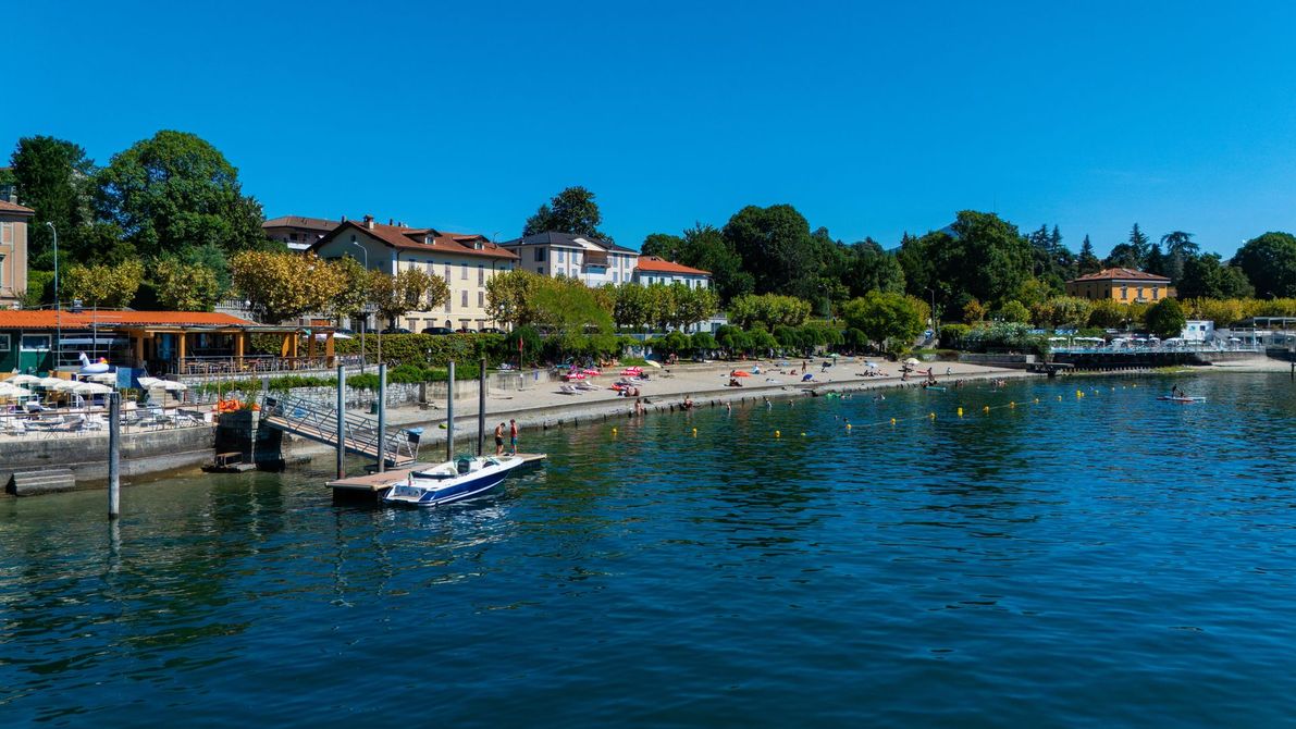 Scena di spiaggia al tramonto; persone che prendono il sole, nuotano; città sullo sfondo, alberi verdi, acqua blu.