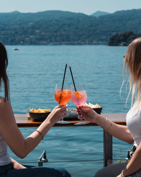 Due mani che brindano a un cocktail Aperol Spritz con un lago sullo sfondo.