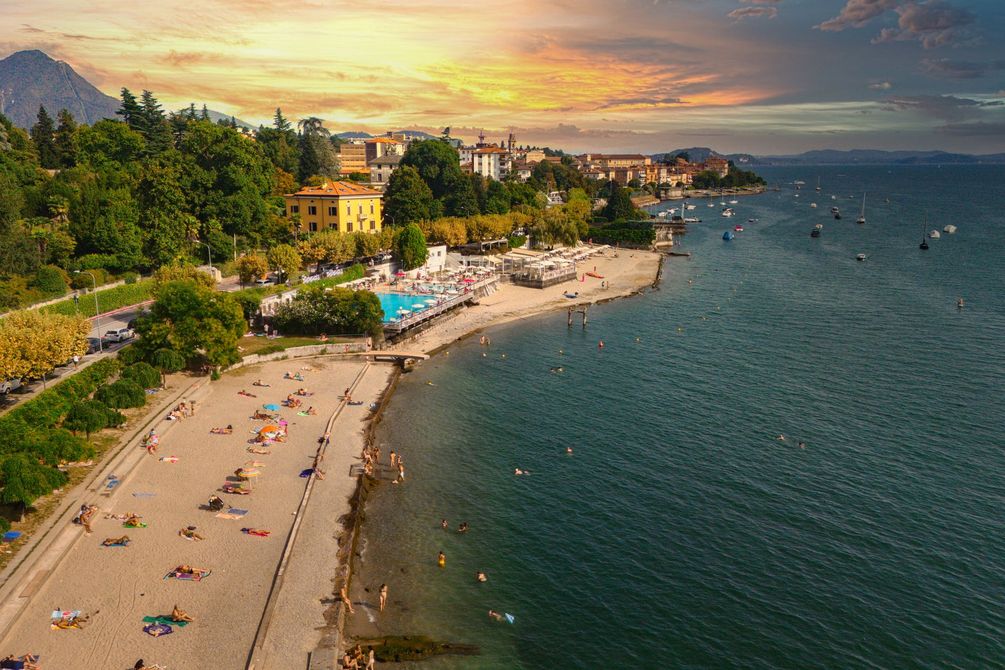 Scena di spiaggia al tramonto; persone che prendono il sole, nuotano; città sullo sfondo, alberi verdi, acqua blu.
