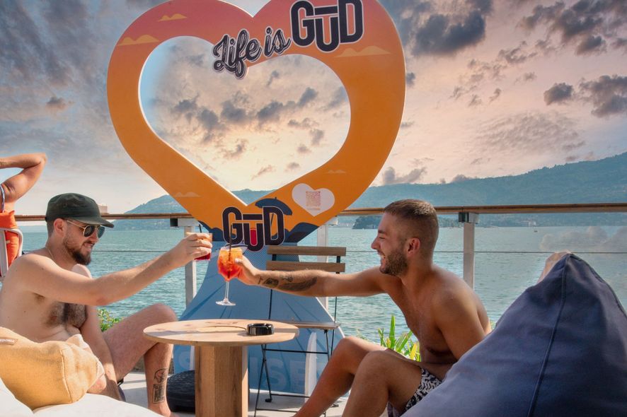 Due uomini a torso nudo brindano su una spiaggia, sotto un cartello a forma di cuore che dice