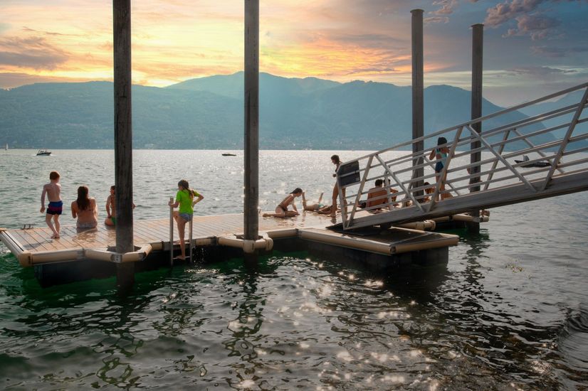 Persone su un molo in un lago al tramonto, con una rampa che conduce verso l'alto e montagne sullo sfondo.