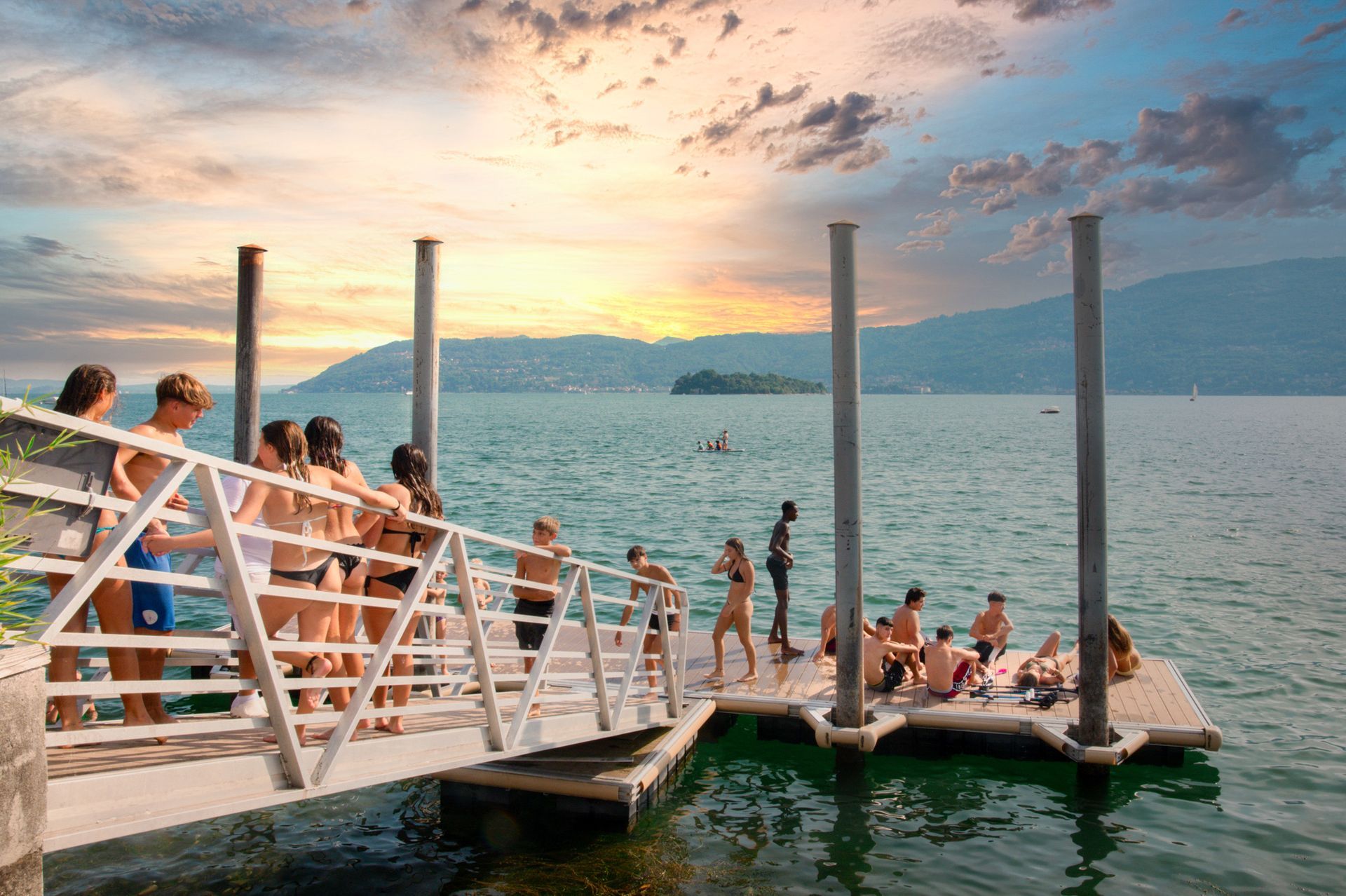 Persone su un molo che nuotano in un lago al tramonto con le montagne sullo sfondo.